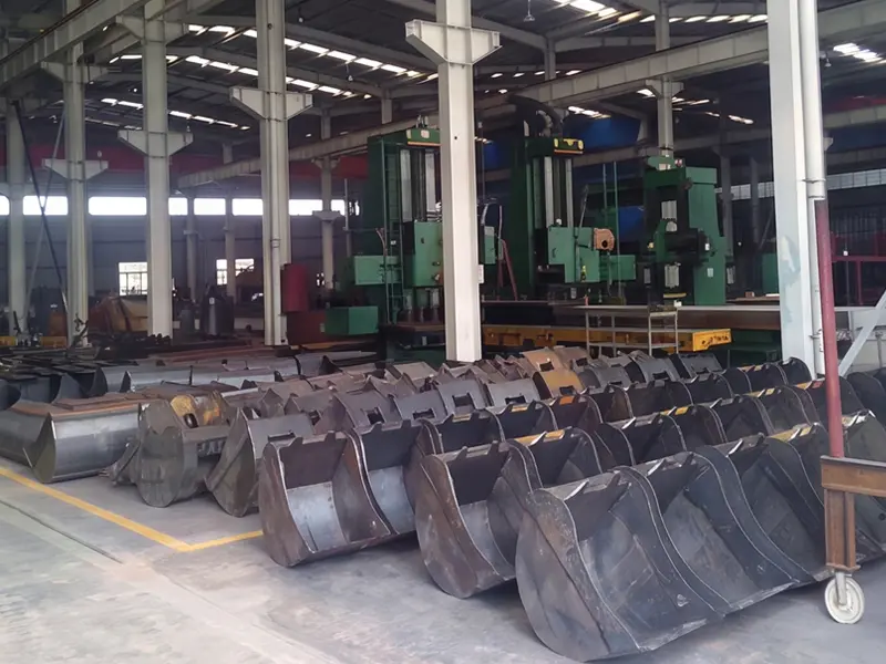 Unpainted excavator bucket structures arranged on a workshop floor with manufacturing equipment behind them
