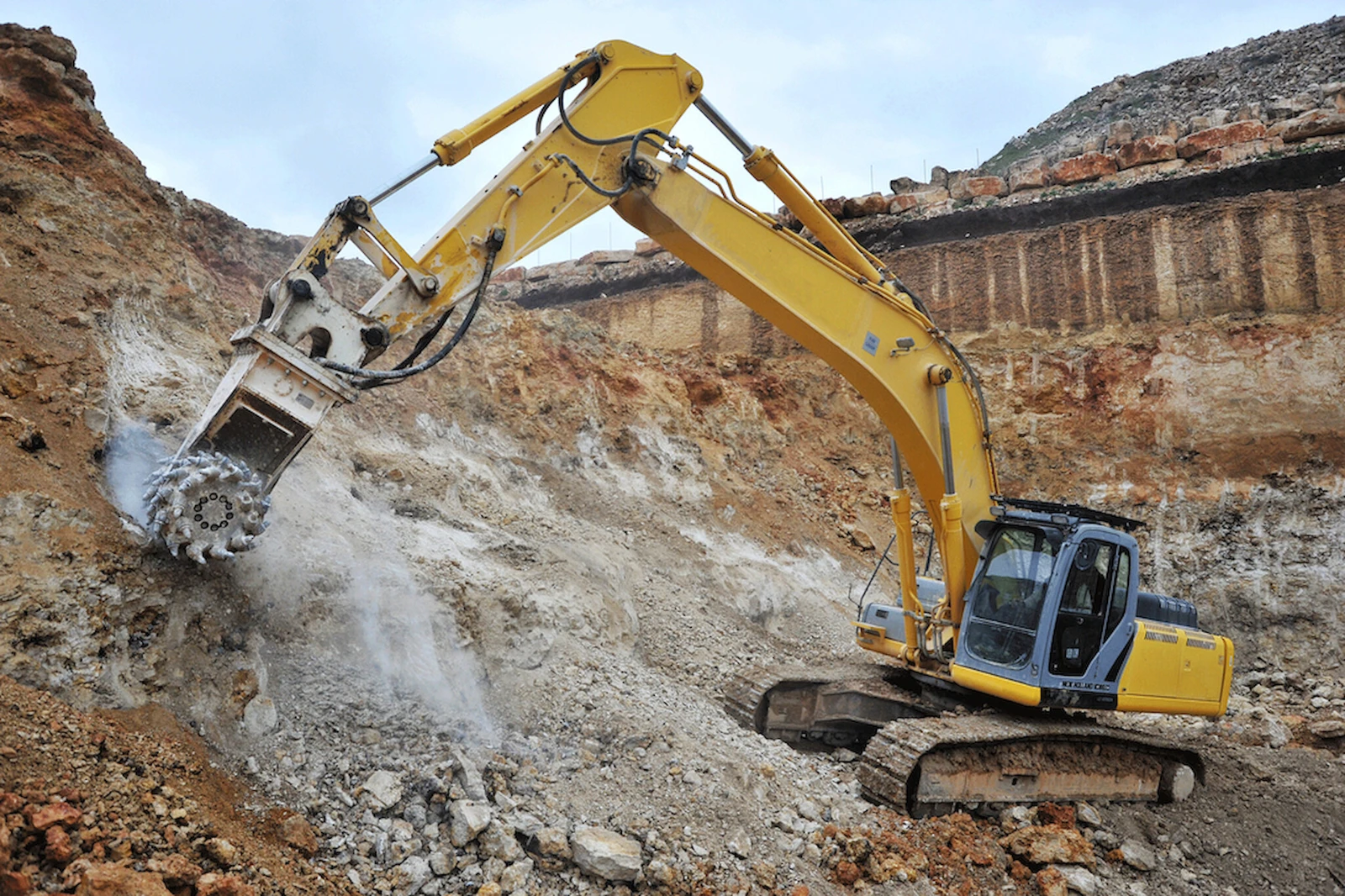 Excavator-mounted transverse drum cutter milling a rock face with dust at the cutting point